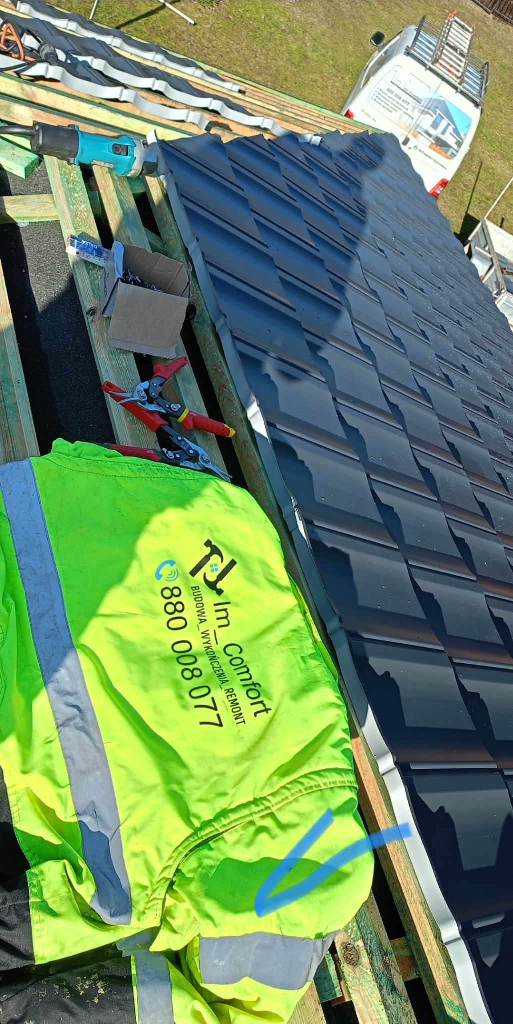 Montaż ciemnoszarej blachodachówki modułowej na drewnianej konstrukcji dachu, widoczne narzędzia dekarskie, odblaskowa kurtka robocza z logo firmy i zaparkowany w pobliżu samochód dostawczy.