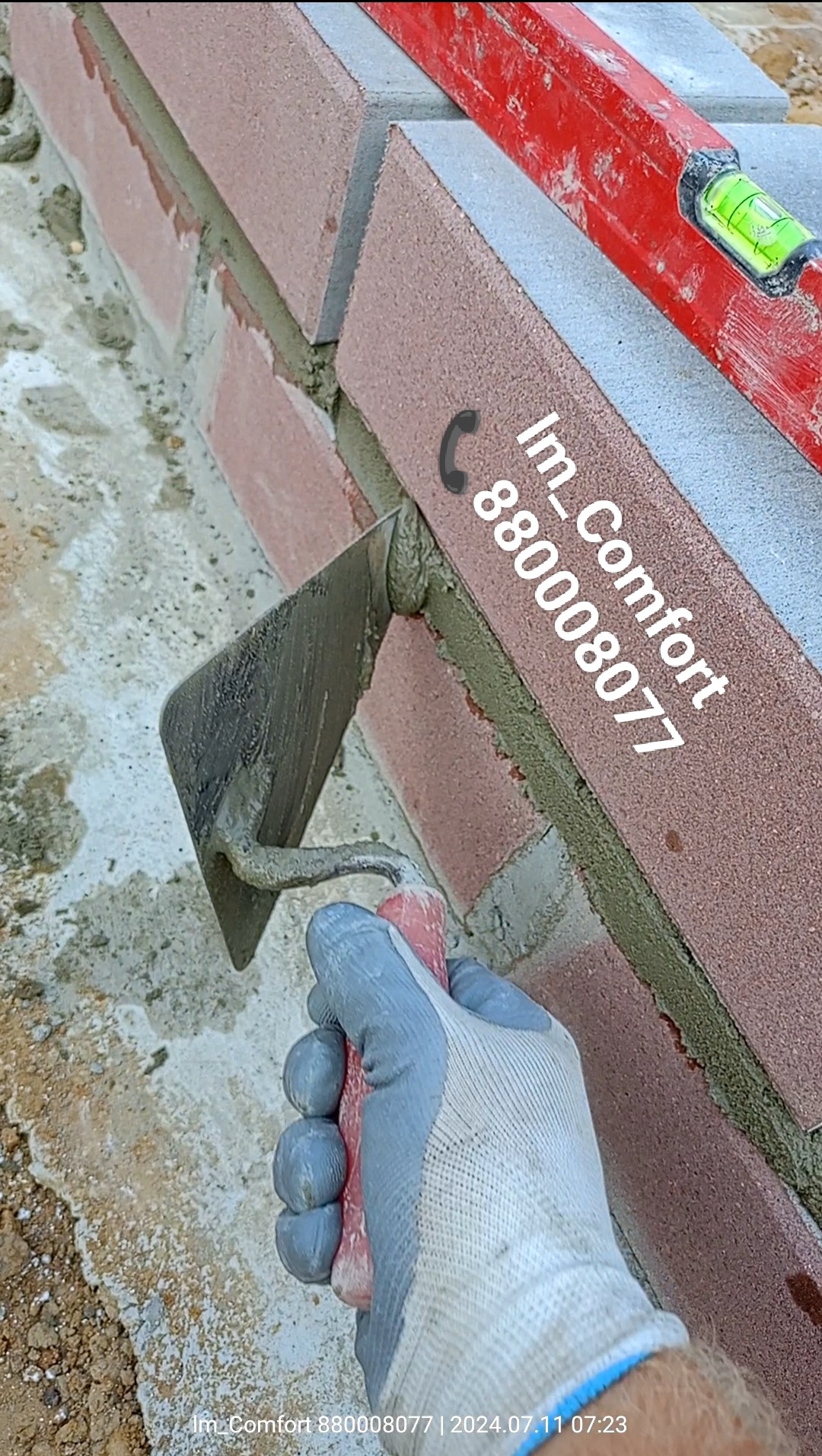 Murowanie ściany z cegły klinkierowej, widoczna kielnia z zaprawą w dłoni w rękawicy oraz poziomica na górze muru.