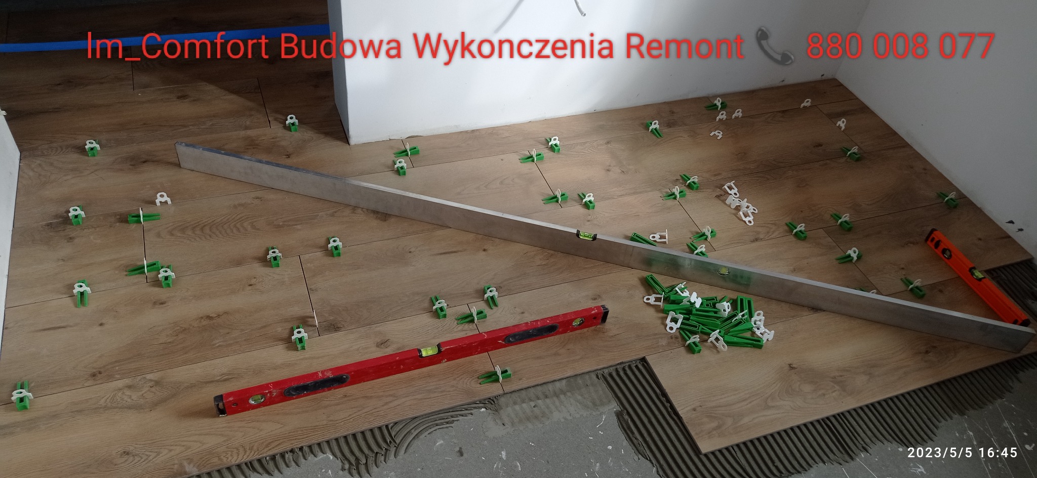 Układanie paneli podłogowych imitujących drewno, z użyciem poziomicy, listwy i zielonych klipsów dystansowych, widoczny klej na podłodze.