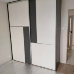 Nowoczesna szafa wnękowa z przesuwnymi drzwiami w kolorze białym i szarym. Widoczny fragment podłogi zabezpieczony folią malarską.