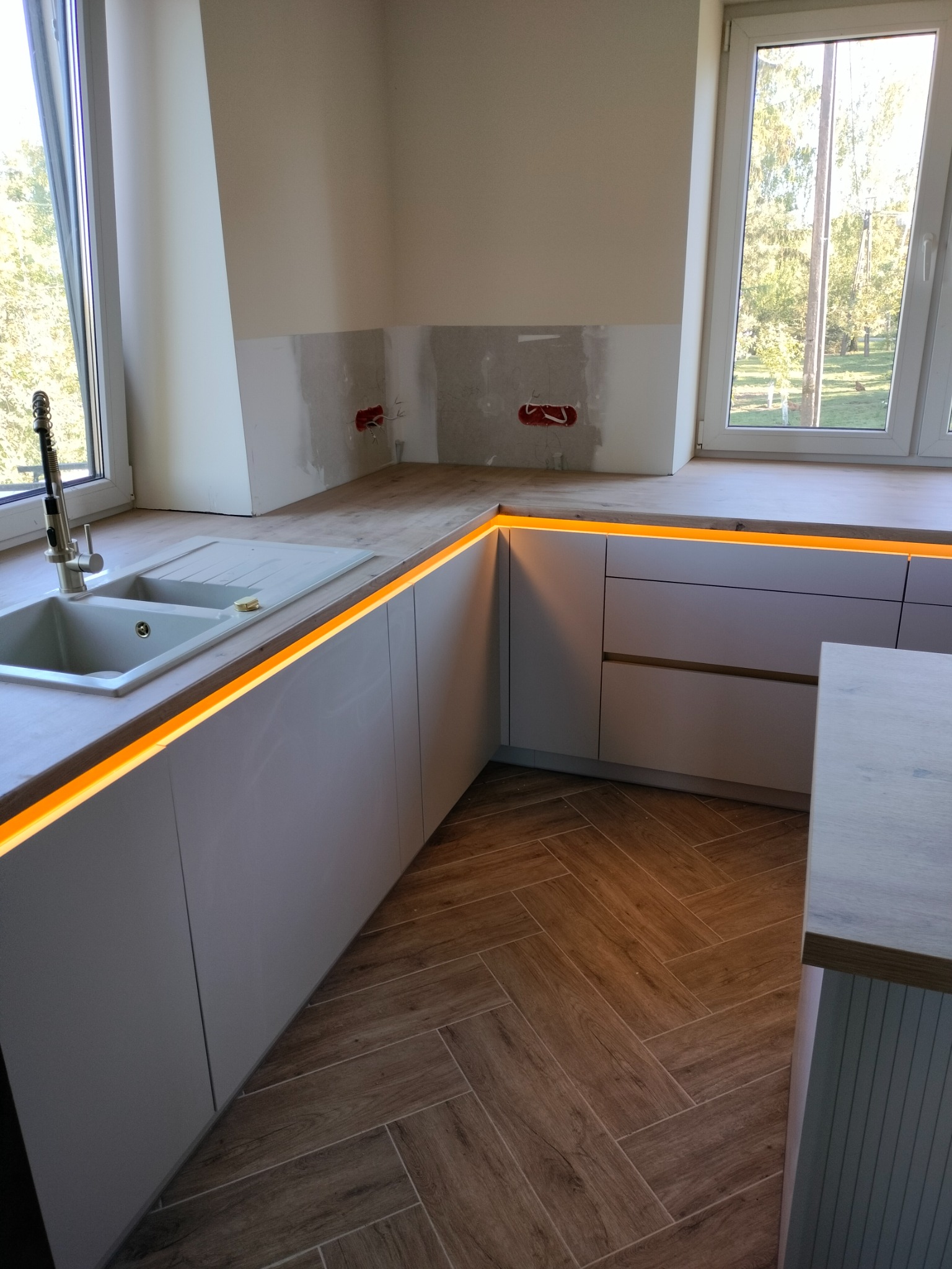 Kuchnia z białymi szafkami, drewnianym blatem i podświetleniem LED. Widok na okno i drewnopodobne płytki na podłodze. Fragment ściany z instalacją elektryczną.