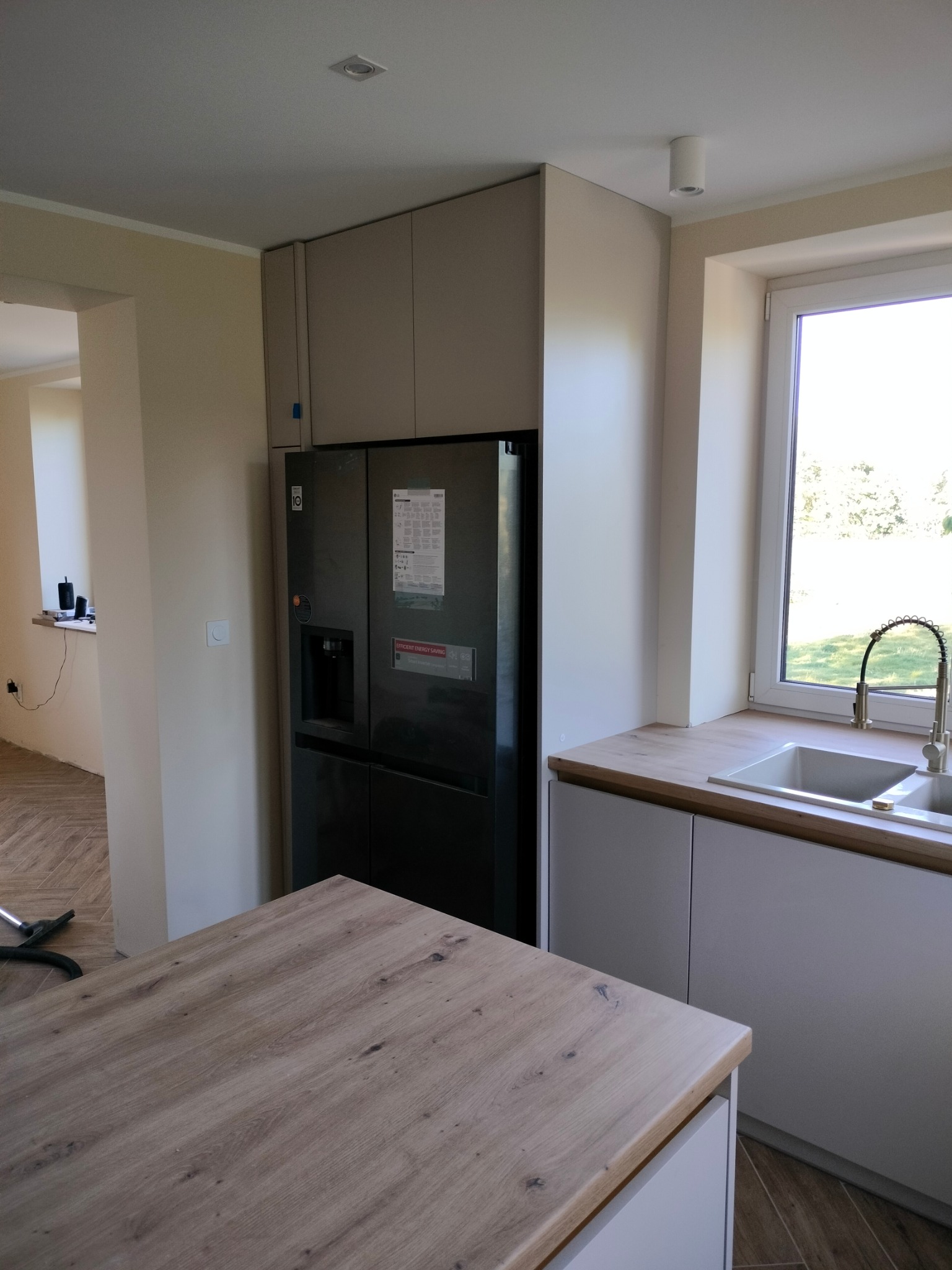 Nowoczesna kuchnia: szare szafki, drewniany blat, czarna lodówka side-by-side. Ujęcie z perspektywy blatu wyspy kuchennej, widok na okno i zlewozmywak.
