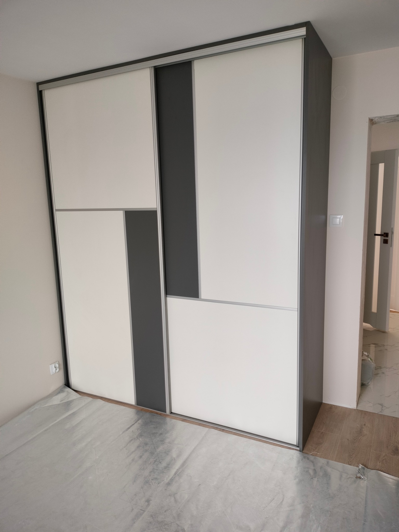 Nowoczesna szafa wnękowa z przesuwnymi drzwiami w kolorze białym i szarym. Widoczny fragment podłogi zabezpieczony folią malarską.