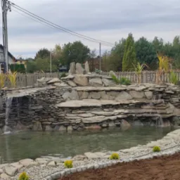 Oczko wodne z kaskadą wodną, wykończone kamieniem naturalnym, otoczone roślinnością ozdobną i obrzeżem z kostki granitowej, widok na ogród i dom w tle.