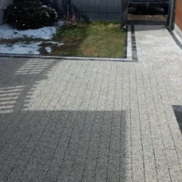 Jasnoszara kostka brukowa ułożona na posesji przydomowej, widoczne fragmenty trawnika z resztkami śniegu i cień rzucany przez konstrukcję.