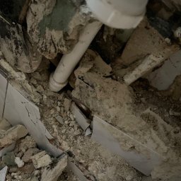 Kostiantyn Pyshniak - Zdemolowany fragment ściany z widocznymi rurami i gruzem, wskazujący na etap prac przygotowawczych do remontu łazienki.