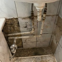 Kostiantyn Pyshniak - Odkryte rury pod zlewem w łazience podczas remontu, widoczne uszkodzone płytki i resztki gruzu. Stare, skorodowane rury wymagają wymiany.
