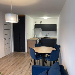 Kostiantyn Pyshniak - Wnętrze mieszkania po remoncie: okrągły stół z czterema granatowymi krzesłami, widoczna kuchnia z czarnymi szafkami i drewnianym blatem, jasna podłoga i sofa w odcieniach granatu.