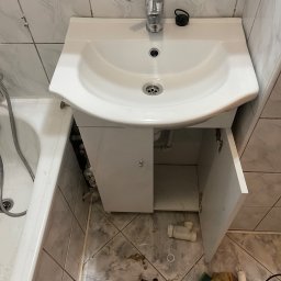 Kostiantyn Pyshniak - Umywalka z szafką w trakcie montażu, otwarte drzwiczki ukazują instalację hydrauliczną, widoczne narzędzia i elementy instalacyjne na podłodze z szarych płytek.