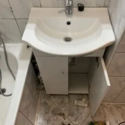 Umywalka z szafką w trakcie montażu, otwarte drzwiczki ukazują instalację hydrauliczną, widoczne narzędzia i elementy instalacyjne na podłodze z szarych płytek.