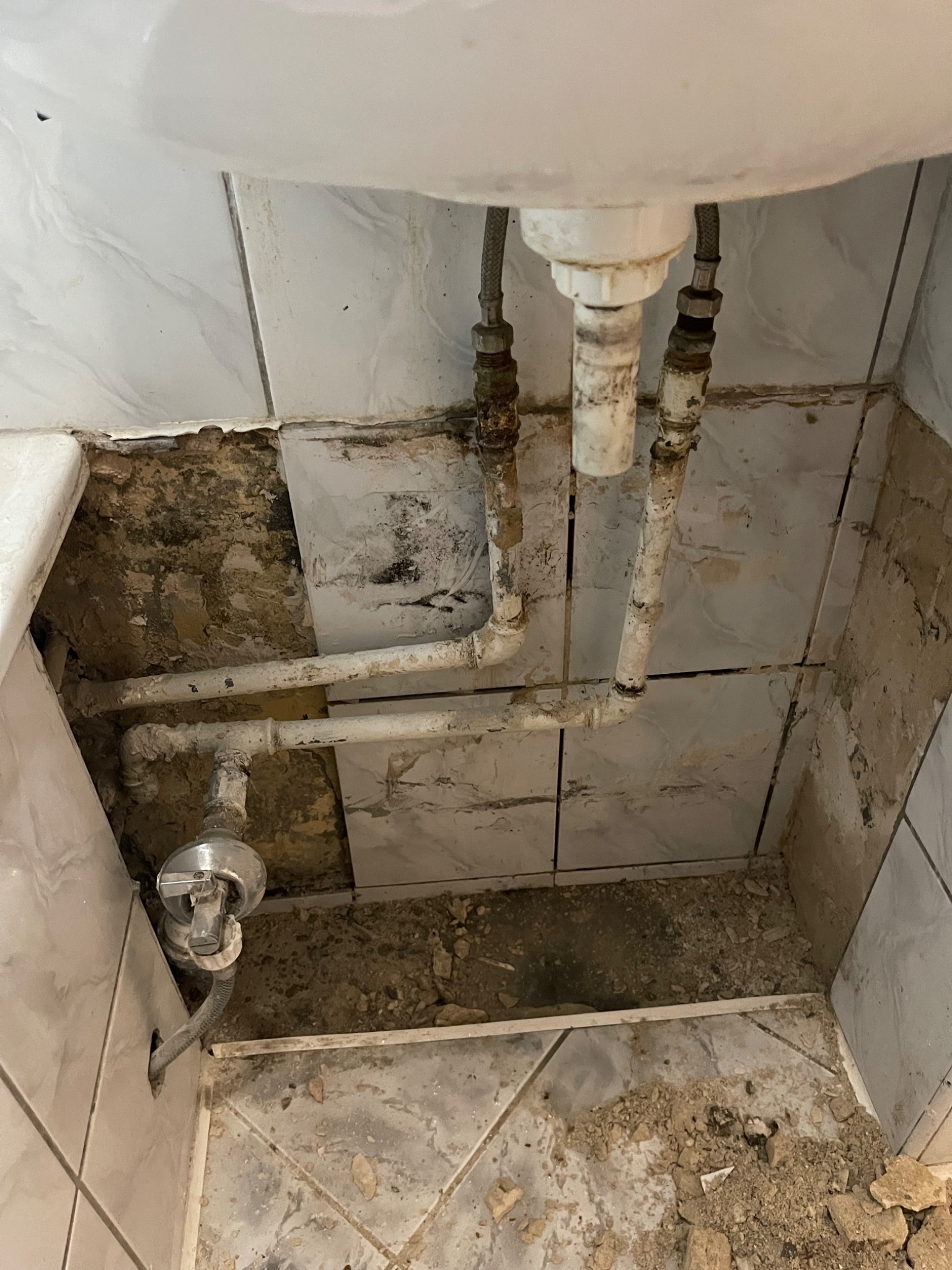 Odkryte rury pod zlewem w łazience podczas remontu, widoczne uszkodzone płytki i resztki gruzu. Stare, skorodowane rury wymagają wymiany.
