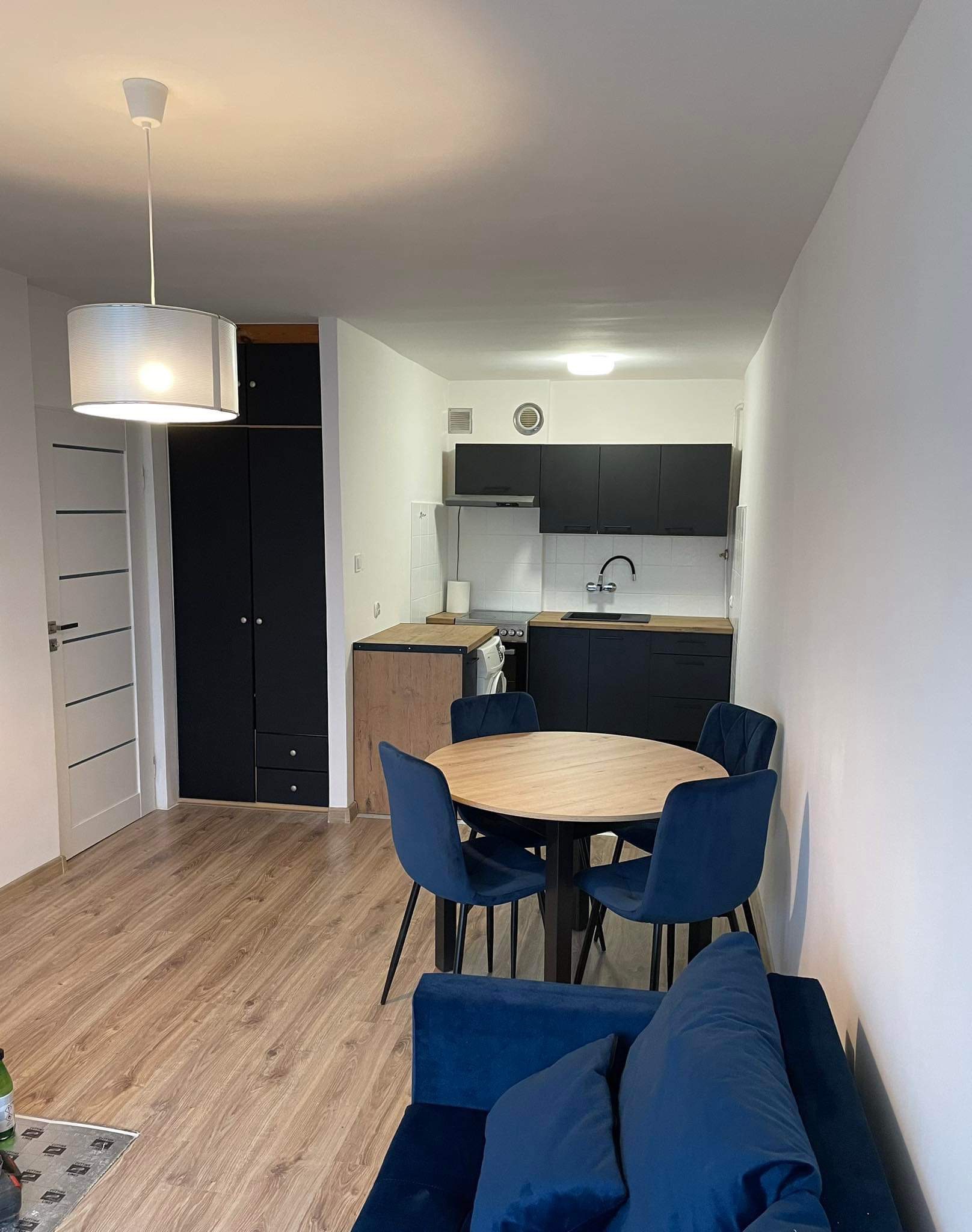Wnętrze mieszkania po remoncie: okrągły stół z czterema granatowymi krzesłami, widoczna kuchnia z czarnymi szafkami i drewnianym blatem, jasna podłoga i sofa w odcieniach granatu.