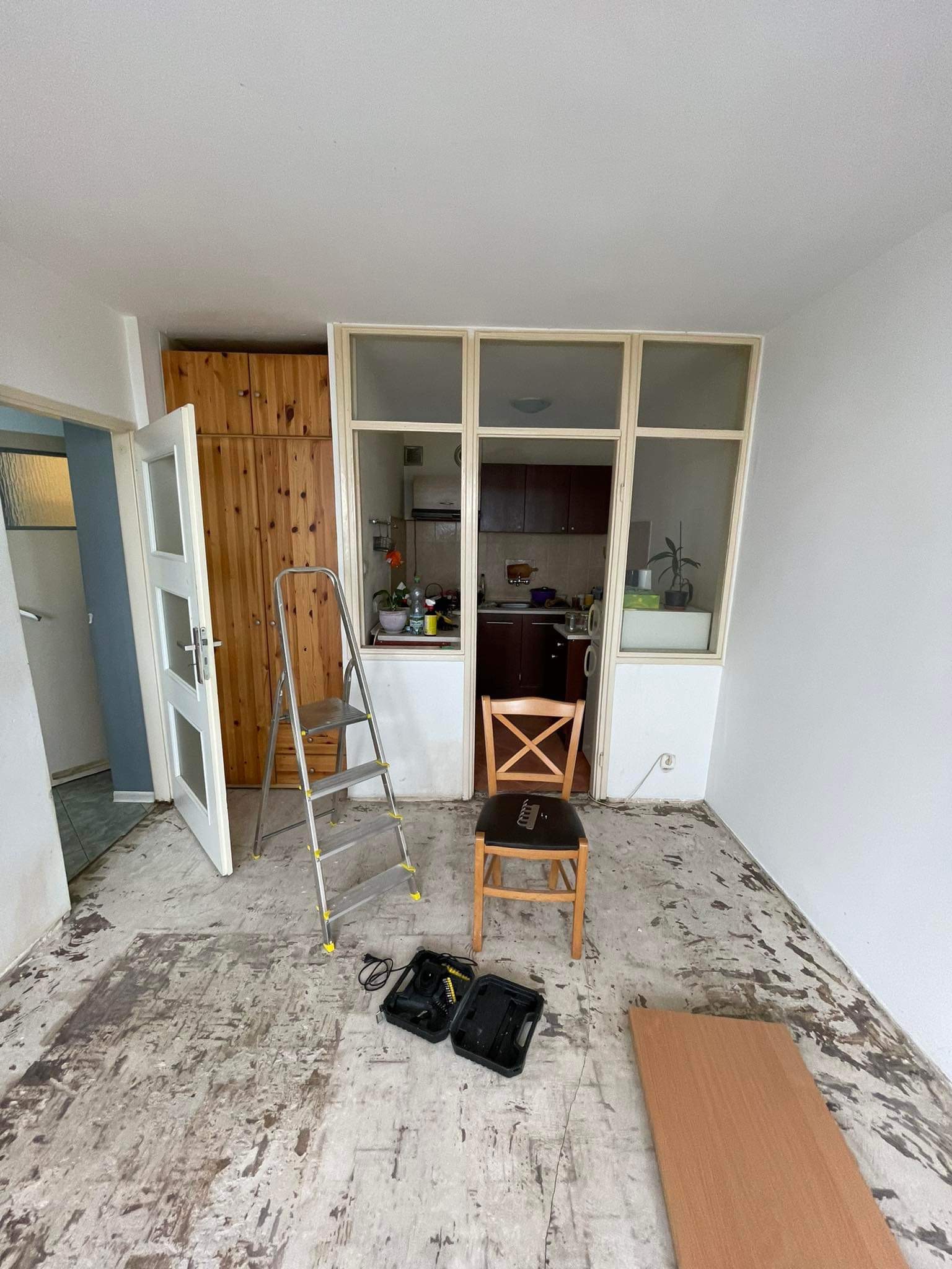 Pokój w trakcie demontażu podłogi, widoczne ślady po usuniętej wykładzinie, drabina, krzesło i skrzynka z narzędziami.