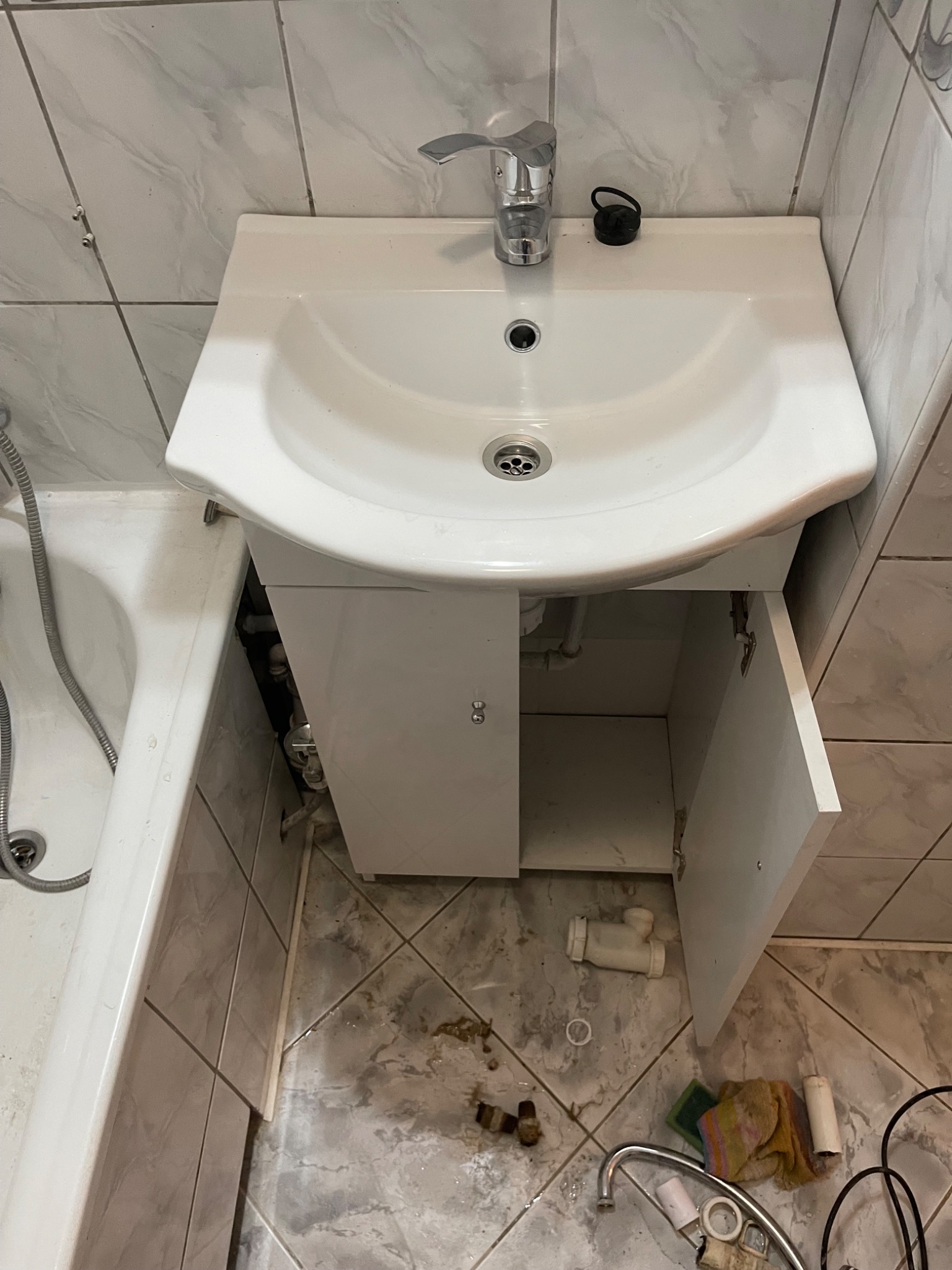 Umywalka z szafką w trakcie montażu, otwarte drzwiczki ukazują instalację hydrauliczną, widoczne narzędzia i elementy instalacyjne na podłodze z szarych płytek.