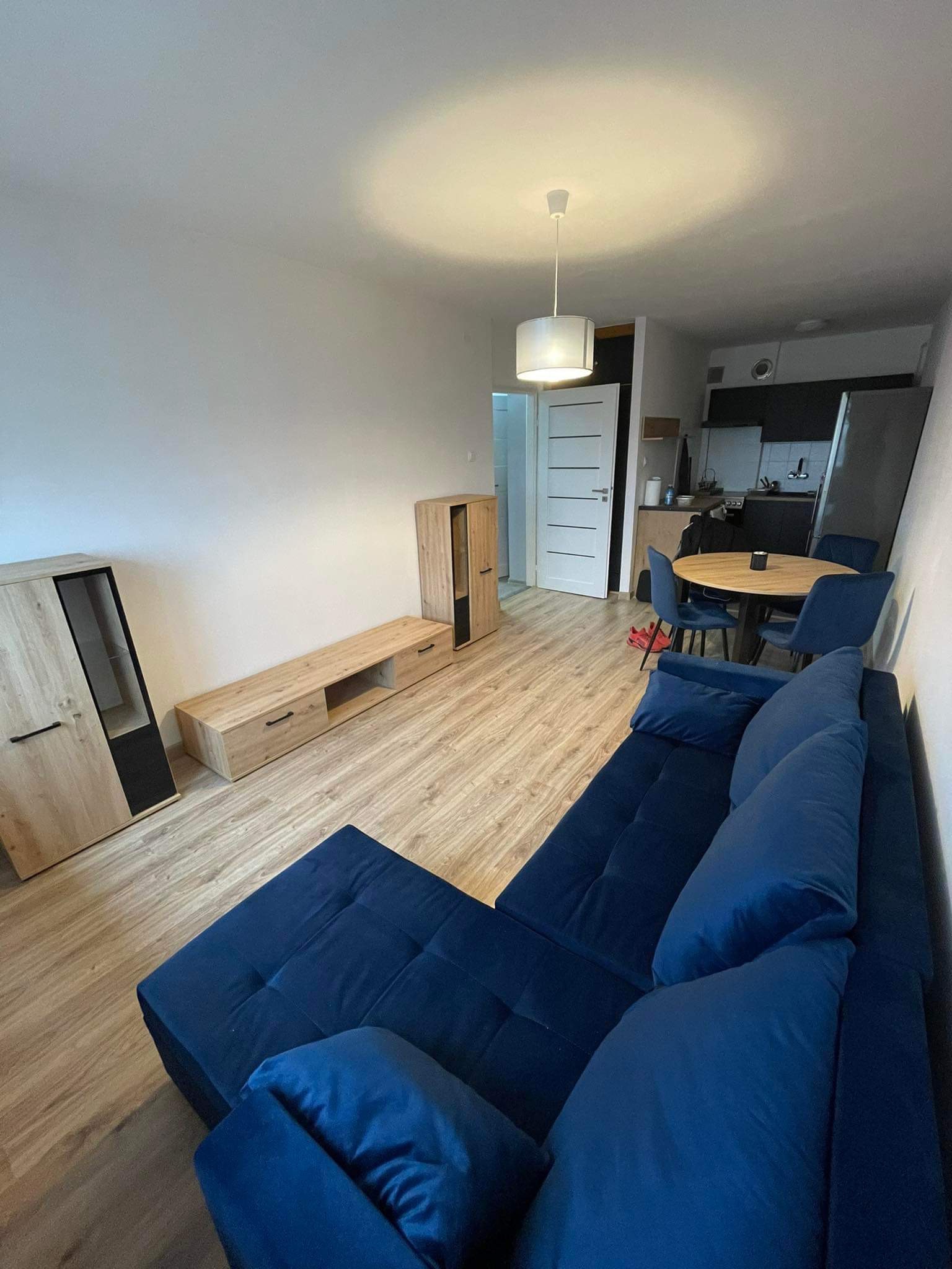 Jasne wnętrze mieszkania po remoncie: granatowy narożnik, drewniane meble, okrągły stół z krzesłami i widoczny fragment kuchni.