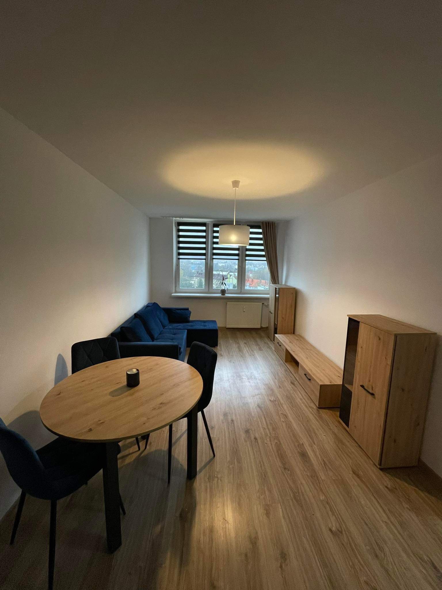 Nowoczesne wnętrze salonu z drewnianą podłogą, okrągłym stołem z czarnymi krzesłami, granatową sofą narożną i meblami w odcieniach drewna, oświetlone wiszącą lampą z abażurem.