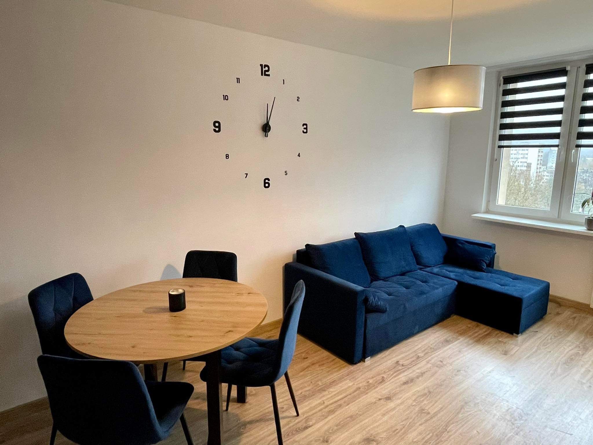 Jasne wnętrze pokoju dziennego z okrągłym stołem, czterema krzesłami, granatową sofą narożną i naklejonym na ścianie zegarem.