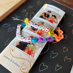 NEUMANN -Alicja Gołuchowska-Neumann - Pamiątkowe zdjęcia pary Joanna i Łukasz z datą 06.07.2024, ozdobione bransoletką z kolorowych koralików z uśmiechniętymi buziami i literami układającymi imię 'ASIA', leżące na czarnym tle w serduszka.