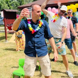 NEUMANN -Alicja Gołuchowska-Neumann - Uczestnicy imprezy integracyjnej w hawajskich wieńcach, jeden z mężczyzn trzyma plastikowy kubek z napojem, w tle zielone krzesło i dekoracje.