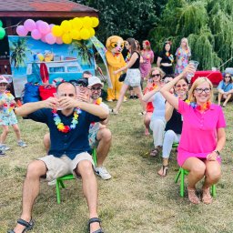 NEUMANN -Alicja Gołuchowska-Neumann - Uczestnicy imprezy plenerowej w hawajskim stylu siedzą na zielonych krzesłach, trzymając plastikowe kubki, w tle dekoracje z balonów, dmuchany autobus i osoba w kostiumie zwierzęcia.