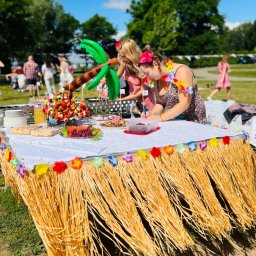 NEUMANN -Alicja Gołuchowska-Neumann - Stół udekorowany w stylu hawajskim z owocowymi przekąskami, balonową palmą i osobami w hawajskich wieńcach, przygotowujący się do imprezy plenerowej.