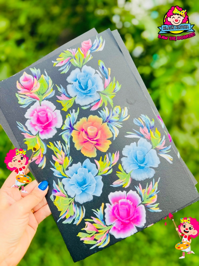 Ręcznie malowane kolorowe kwiaty na czarnym tle, trzymane w dłoni z niebieskim paznokciem, obok naklejka z postacią klauna z paletą farb.