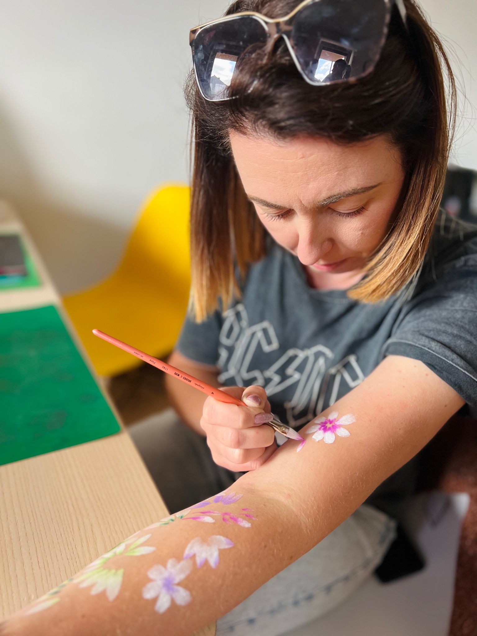 Kobieta maluje kwiaty na ramieniu pędzlem, używając farb do ciała, w tle żółte krzesło.