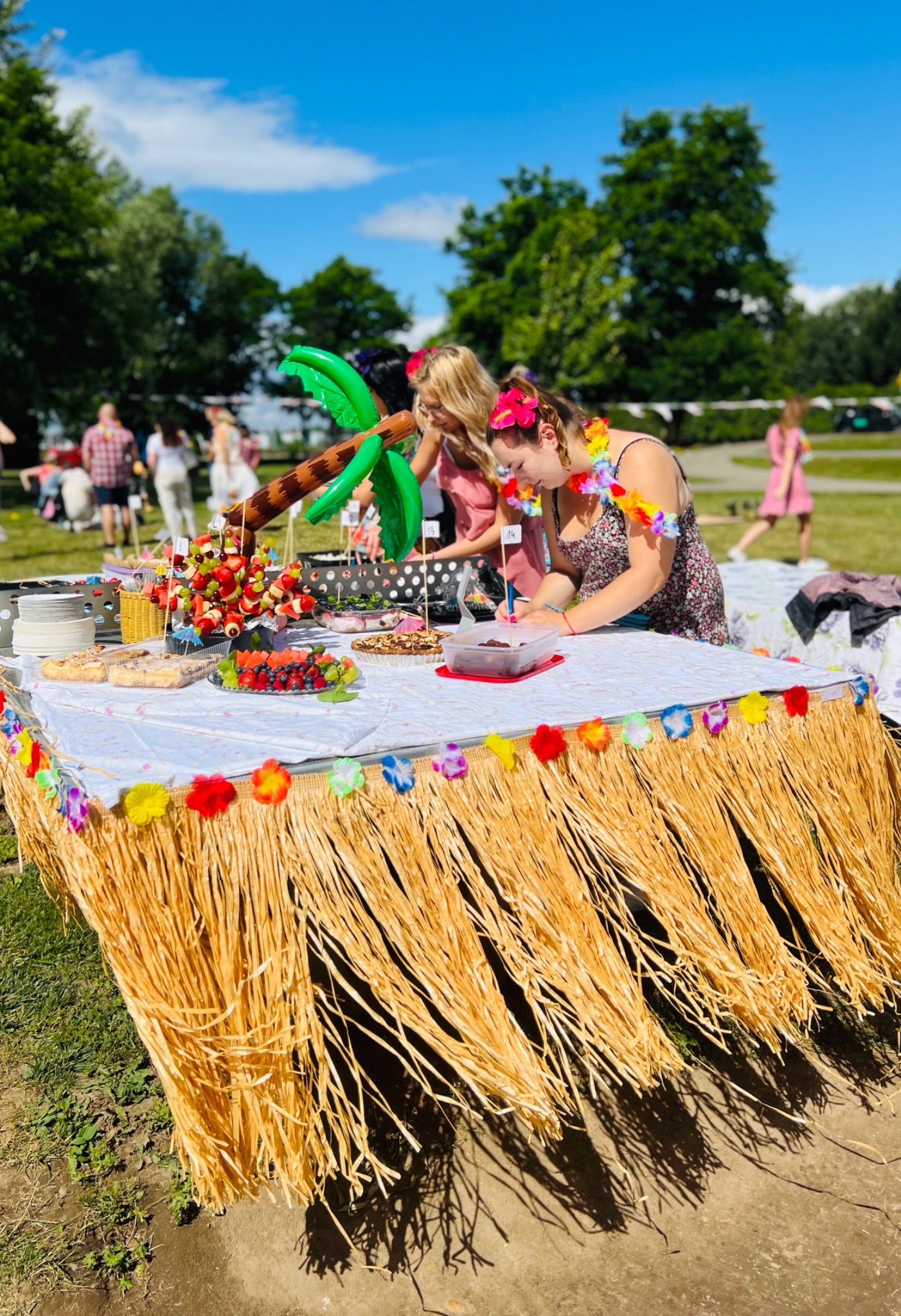 Stół udekorowany w stylu hawajskim z owocowymi przekąskami, balonową palmą i osobami w hawajskich wieńcach, przygotowujący się do imprezy plenerowej.