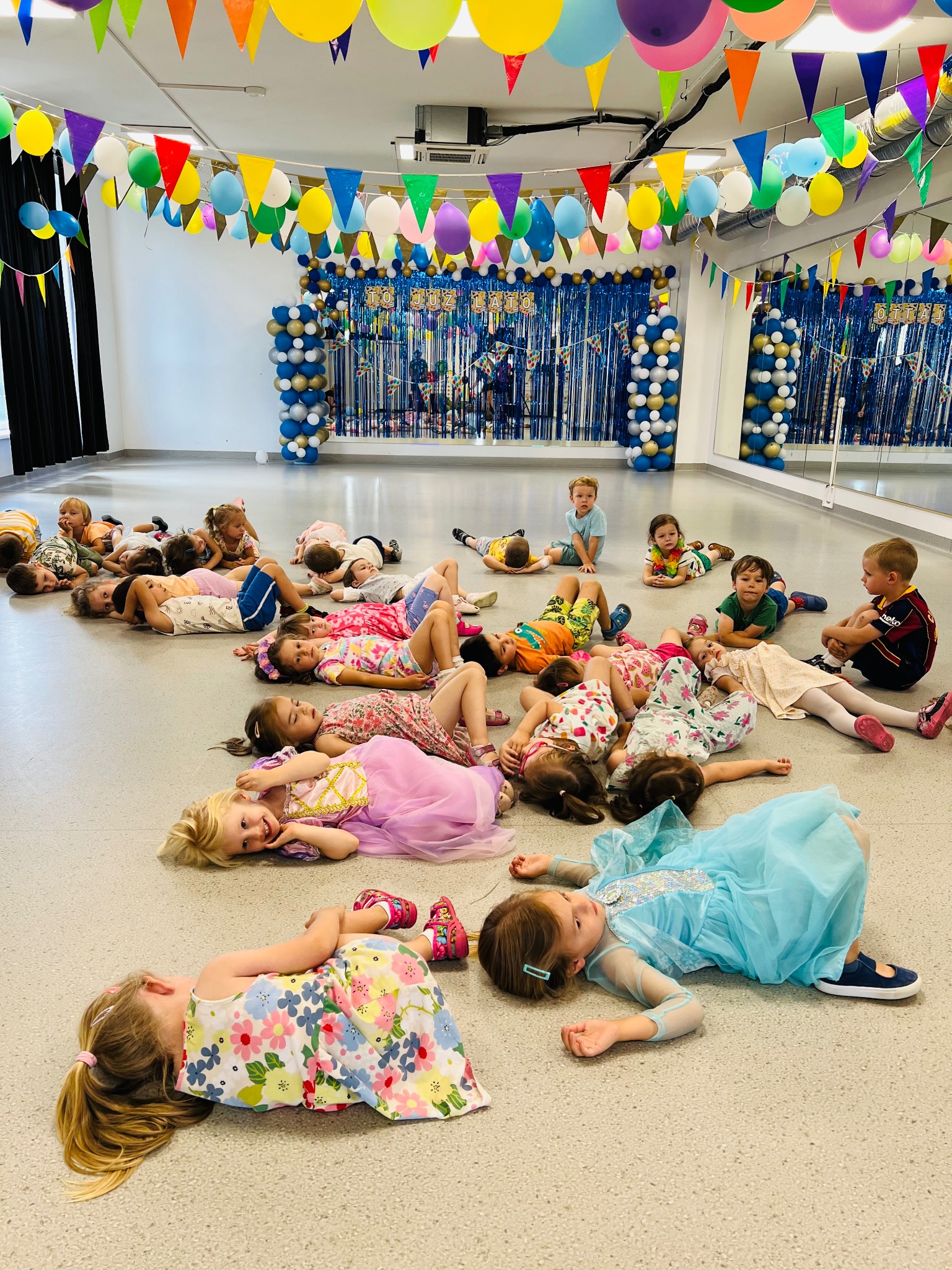 Grupa dzieci leżących na podłodze w udekorowanej sali, część w strojach karnawałowych, w tle dekoracje z balonów i girland.