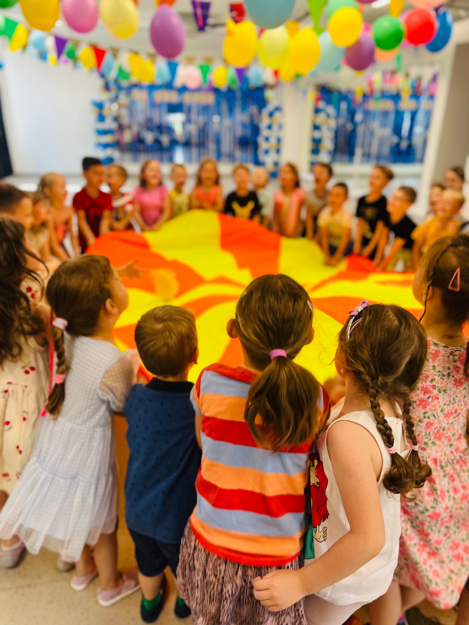 Grupa dzieci otaczająca dużą, okrągłą chustę w odcieniach żółci i pomarańczy, trzymając ją za brzeg, z dekoracjami z balonów i girland w tle.
