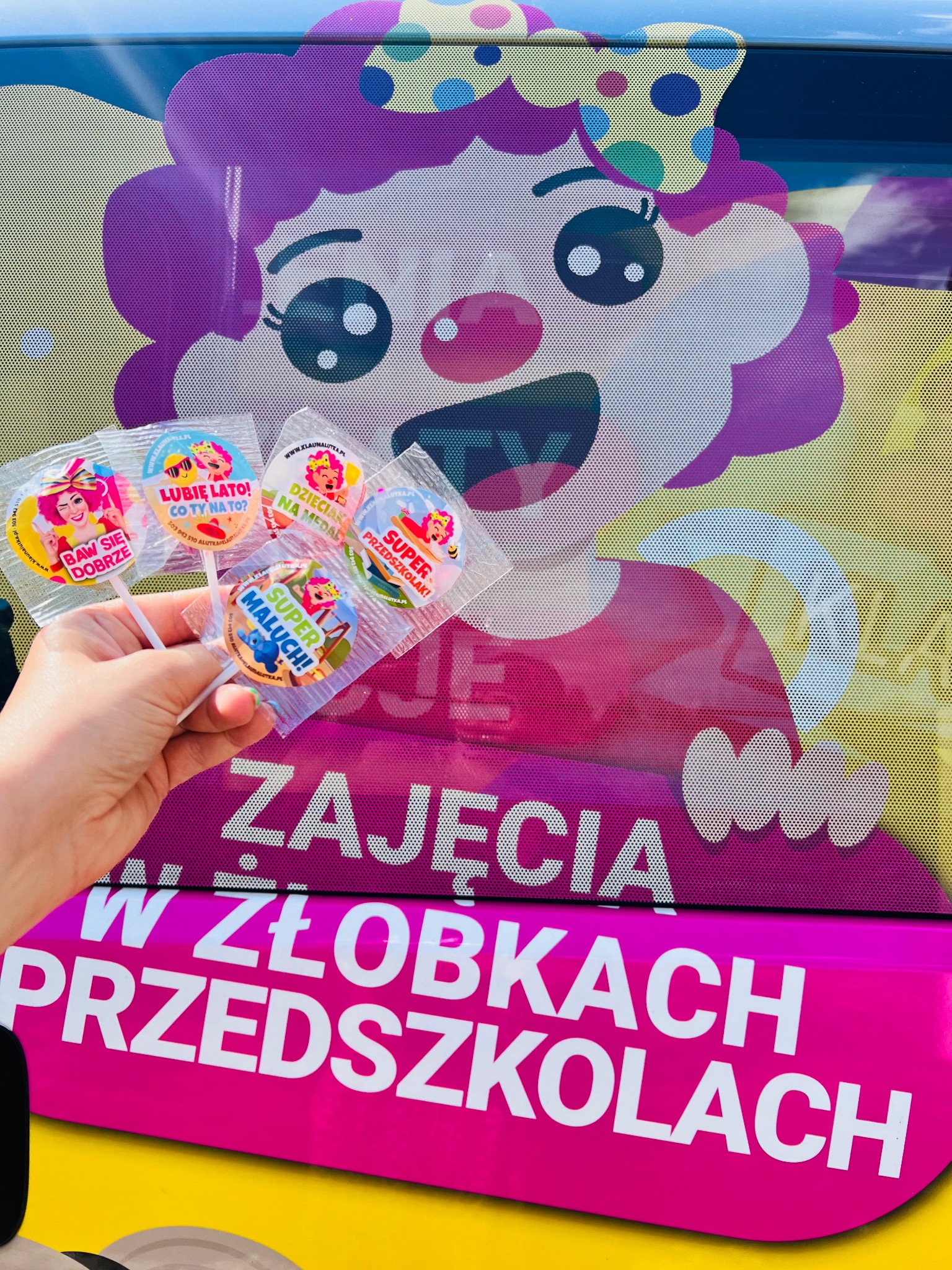 Ręka trzymająca lizaki z wizerunkiem klauna Alutki na tle oklejonego samochodu z napisem 'Zajęcia w żłobkach i przedszkolach' i wizerunkiem klauna.