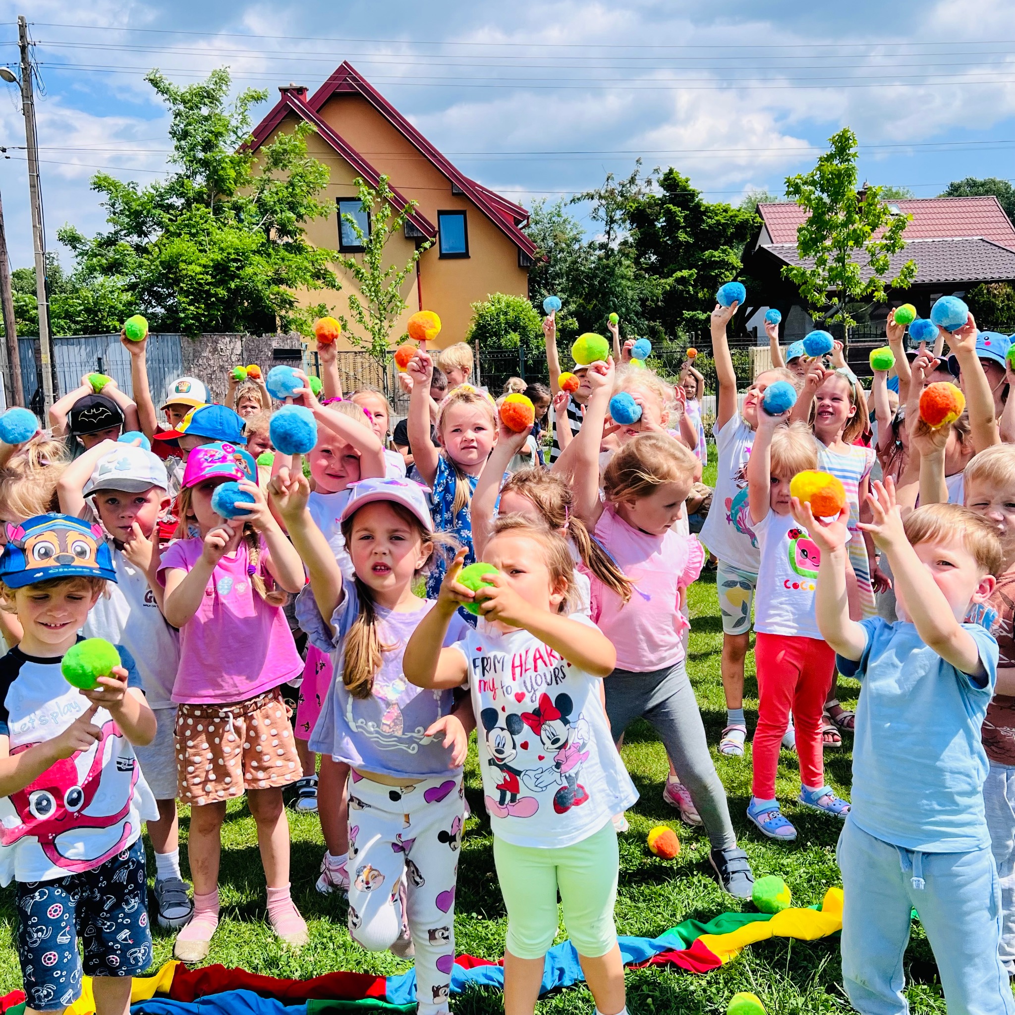 Grupa roześmianych dzieci na trawie trzymających w górze kolorowe, miękkie piłki, w tle dom z czerwonym dachem i zielone drzewa.
