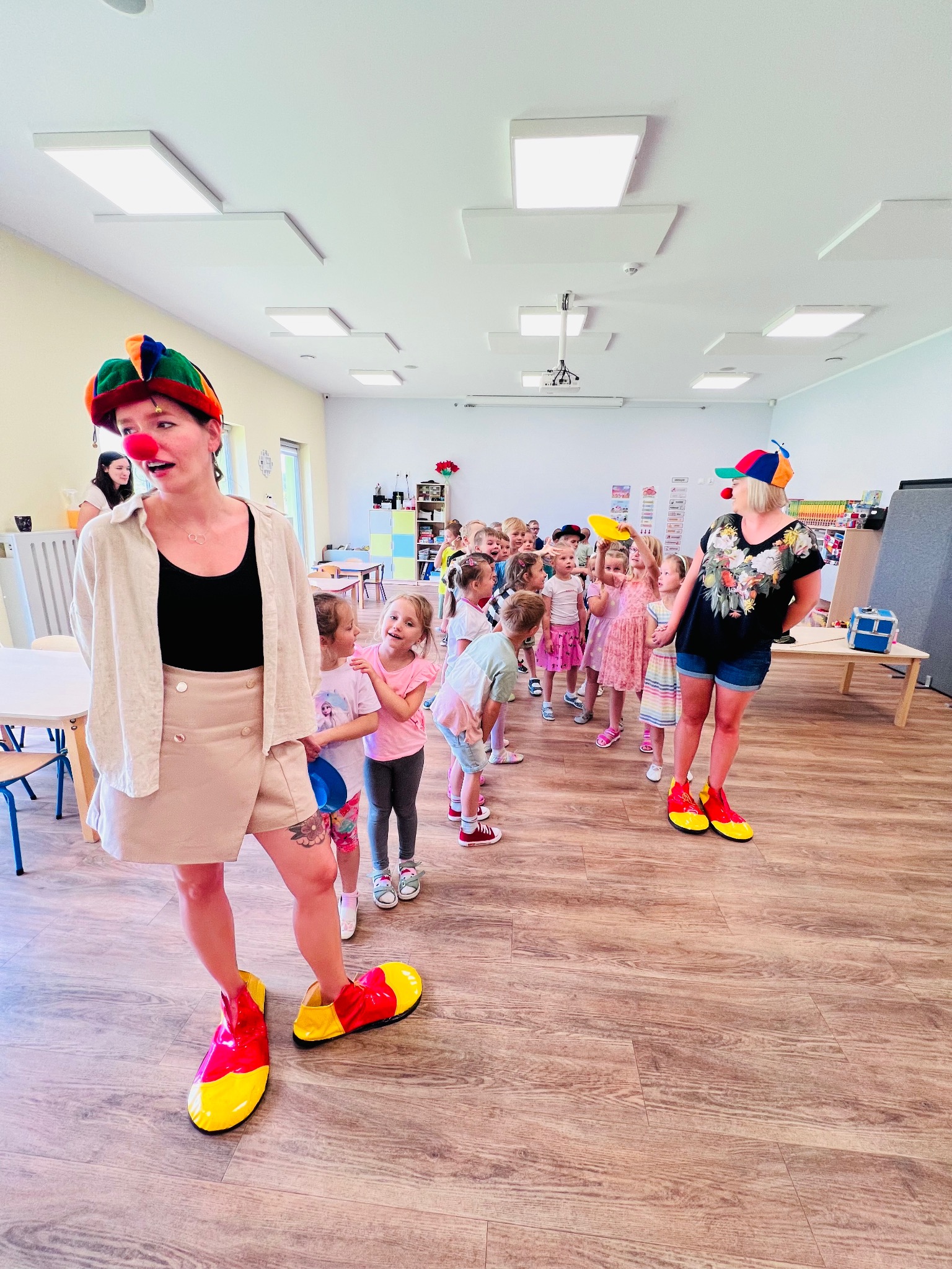 Dwie animatorki w strojach klaunów z czerwonymi nosami i dużymi butami prowadzą grupę dzieci w jasnym pomieszczeniu z drewnianą podłogą. Jedna z animatorek ma na nodze tatuaż z motywem kwiatowym.