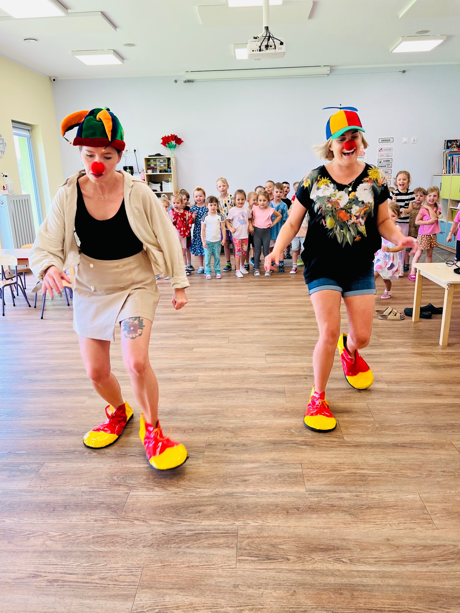 Dwie kobiety ubrane jak klauni w kolorowych strojach i dużych butach tańczą przed grupą dzieci w jasnym pomieszczeniu z drewnianą podłogą.