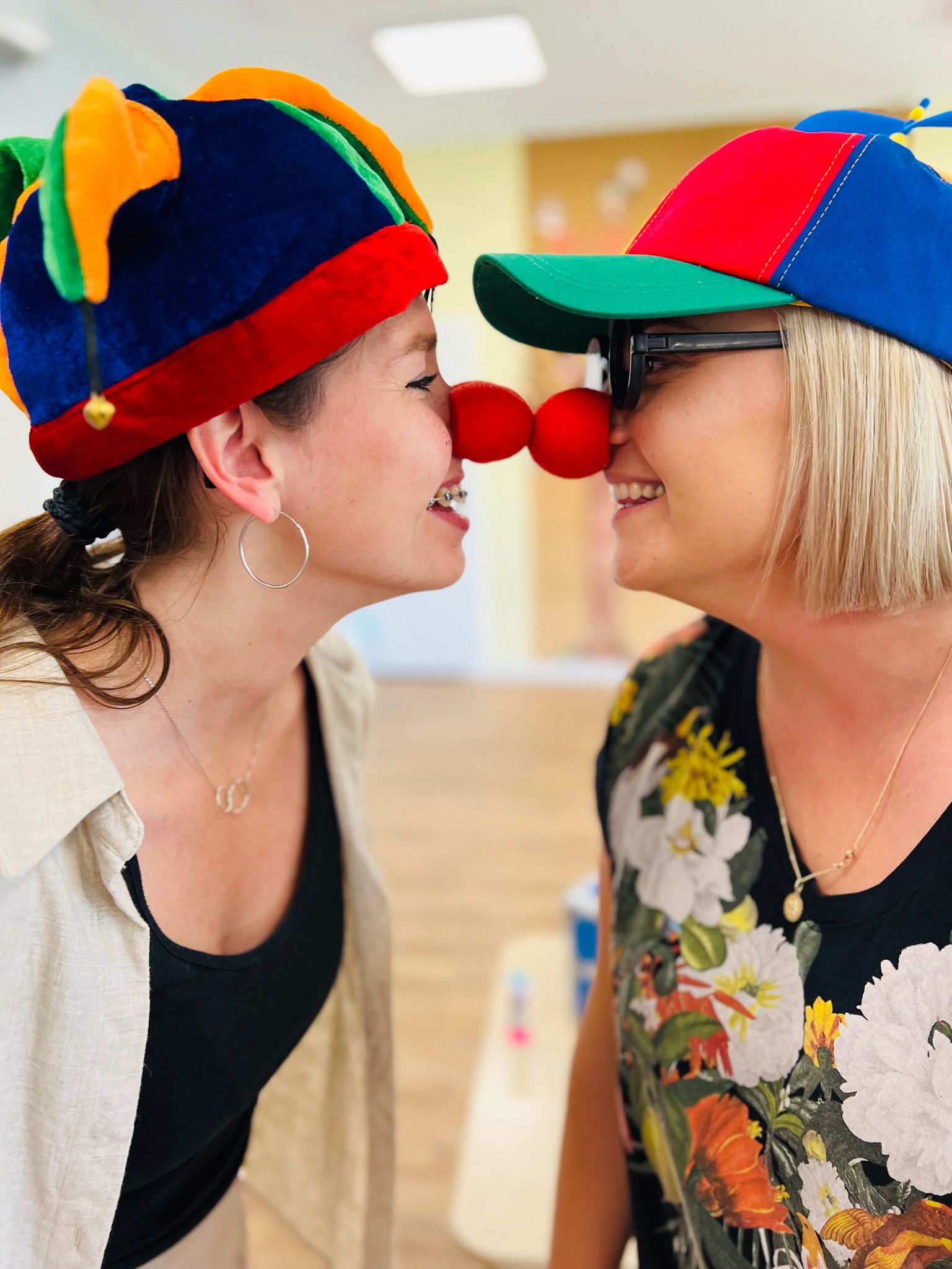Dwie uśmiechnięte kobiety z czerwonymi nosami klauna, w kolorowych kapeluszach, stykają się nosami.