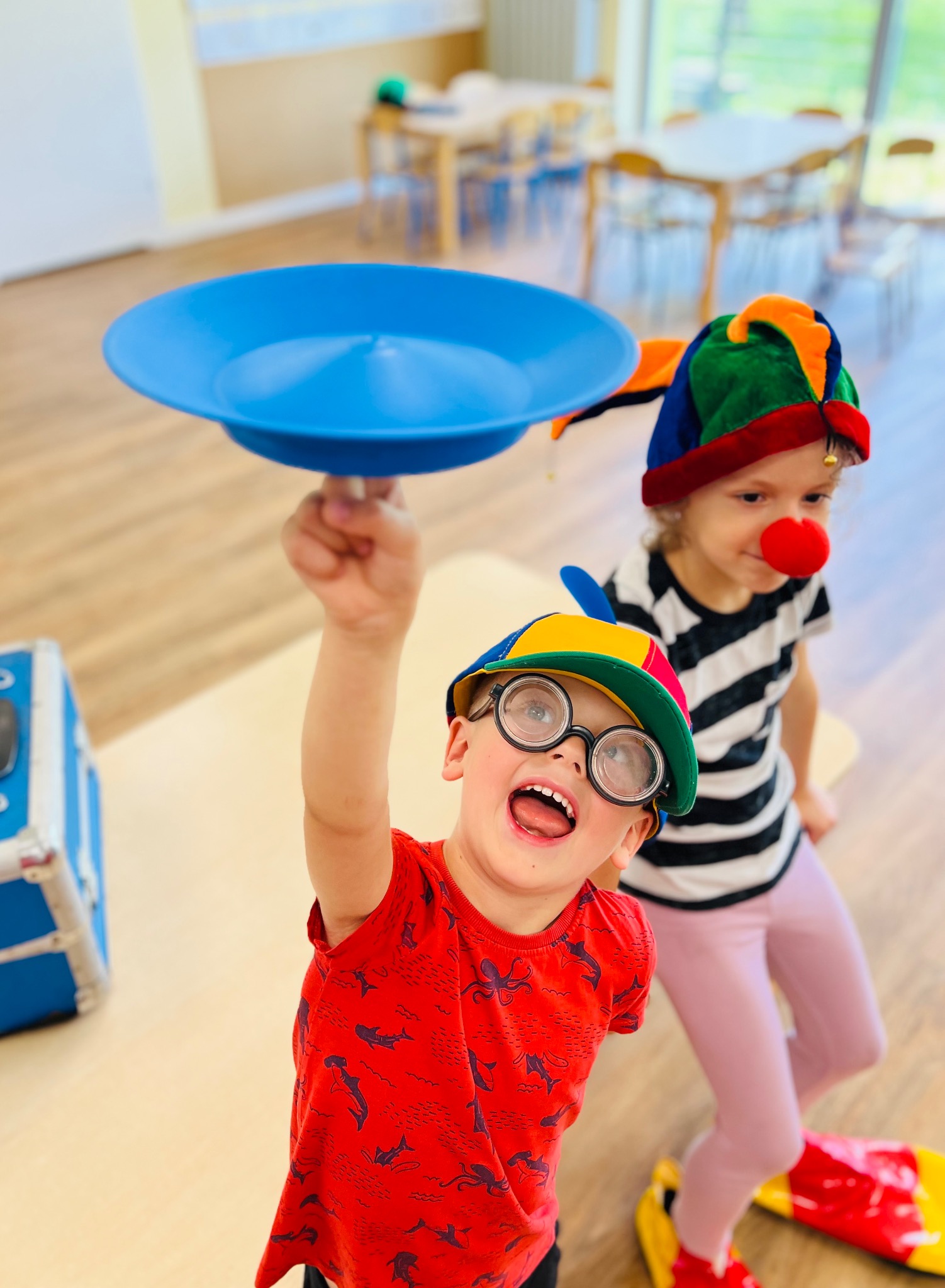 Uśmiechnięty chłopiec w kolorowej czapce i okularach trzyma niebieski talerz na patyku, w tle dziewczynka z czerwonym nosem clowna i w czarno-białej koszulce.