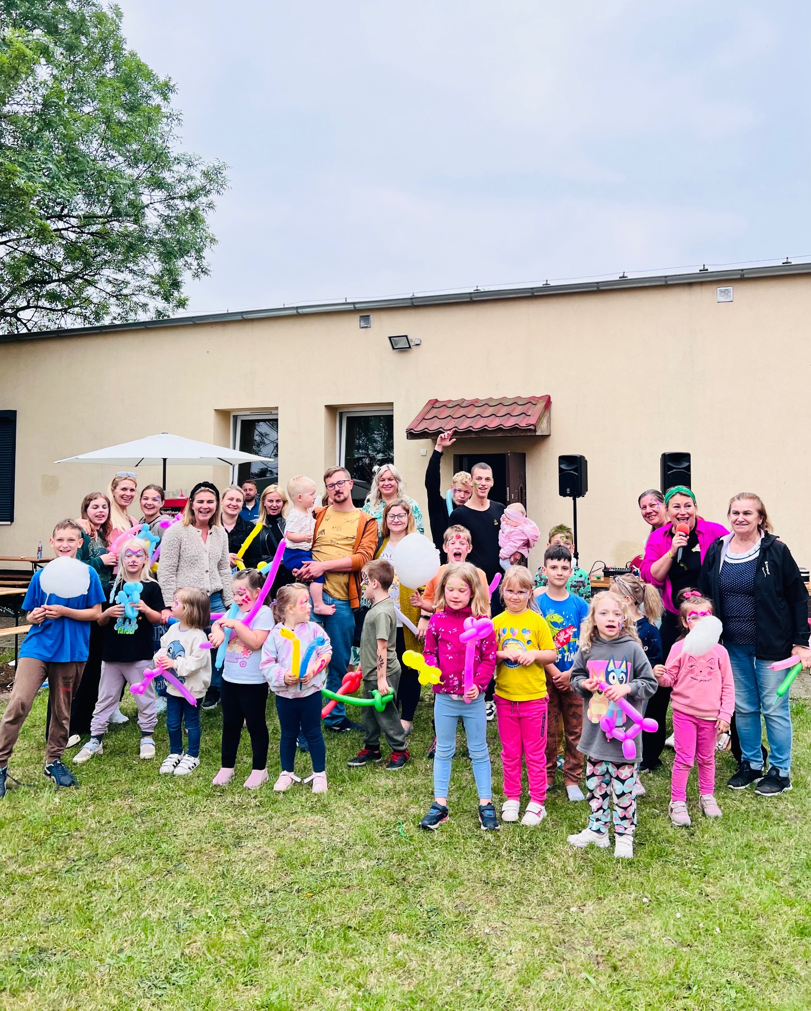 Grupa dzieci i dorosłych pozuje do zdjęcia na trawniku przed budynkiem. Dzieci trzymają balonowe zwierzątka i mają pomalowane twarze. Kobieta z mikrofonem w różowej koszuli i zielonej opasce...