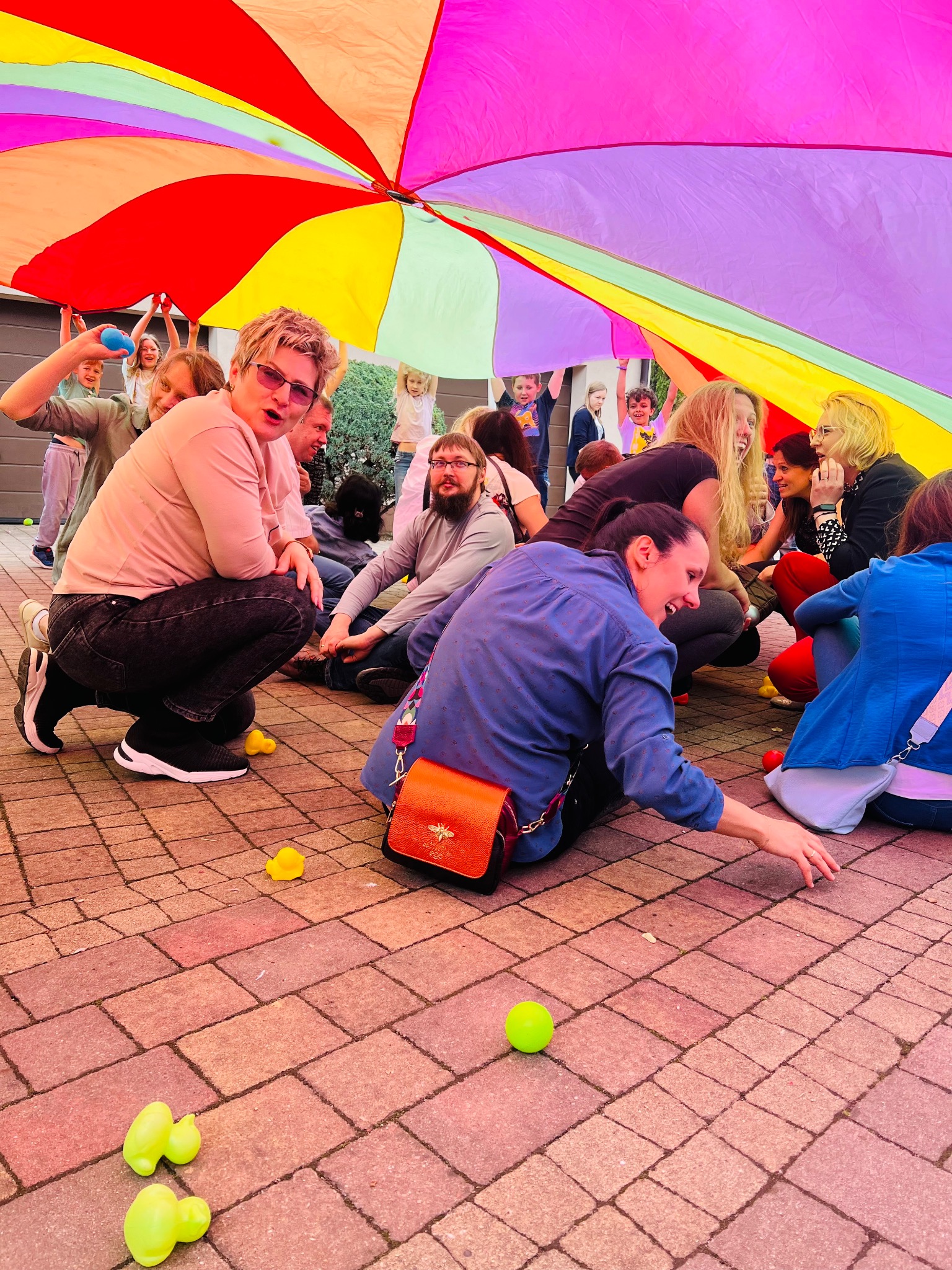 Grupa dorosłych i dzieci siedzących pod kolorową chustą animacyjną na brukowanym placu, z rozrzuconymi żółtymi gumowymi kaczkami i zielonymi piłkami, podczas animacji zorganizowanej na świeżym...