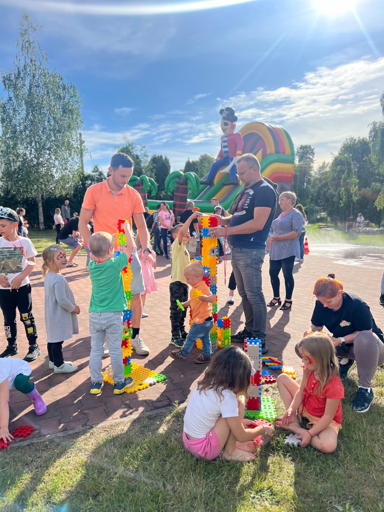 Dzieci budują wieżę z kolorowych, dużych klocków na trawie, pod nadzorem dwóch mężczyzn i kobiety, w tle dmuchany zamek i inne osoby.