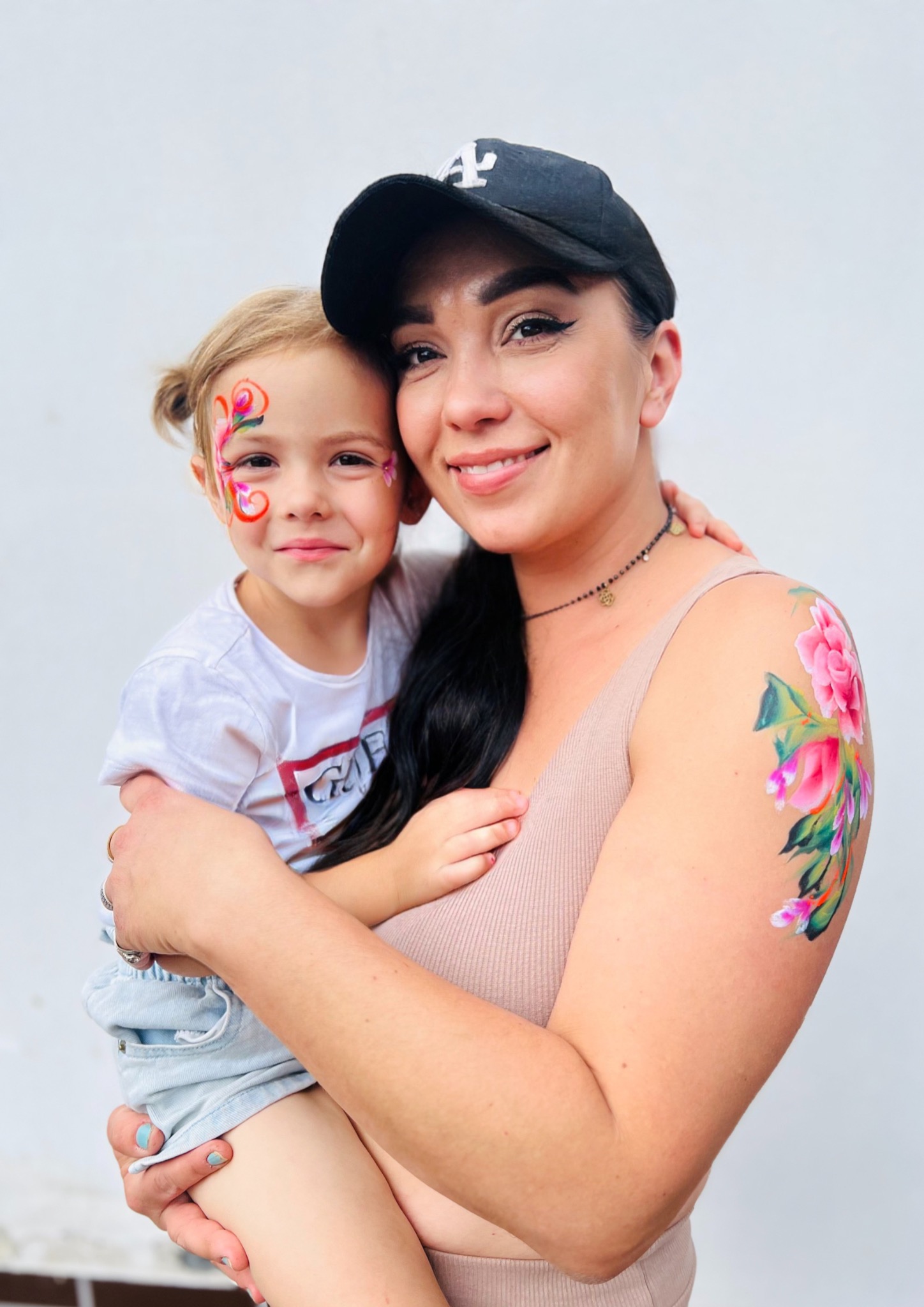 Uśmiechnięta kobieta w czapce trzyma na rękach dziewczynkę z pomalowaną twarzą w motyle. Kobieta ma tatuaż z kwiatami na ramieniu.