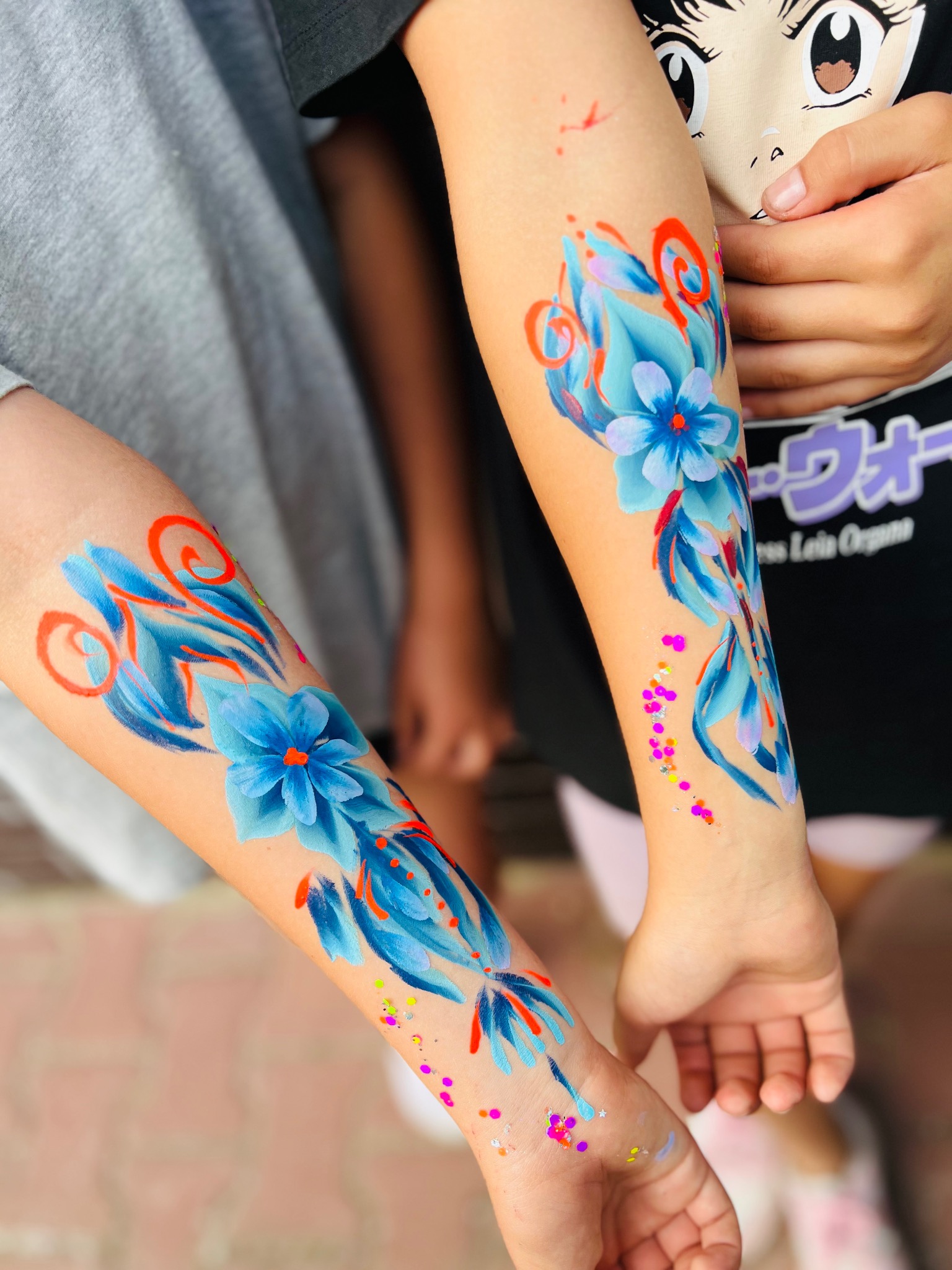 Dwa przedramiona dzieci z pomalowanymi niebieskimi kwiatami z pomarańczowymi akcentami i brokatem, widoczne na tle chodnika i koszulki z motywem anime.