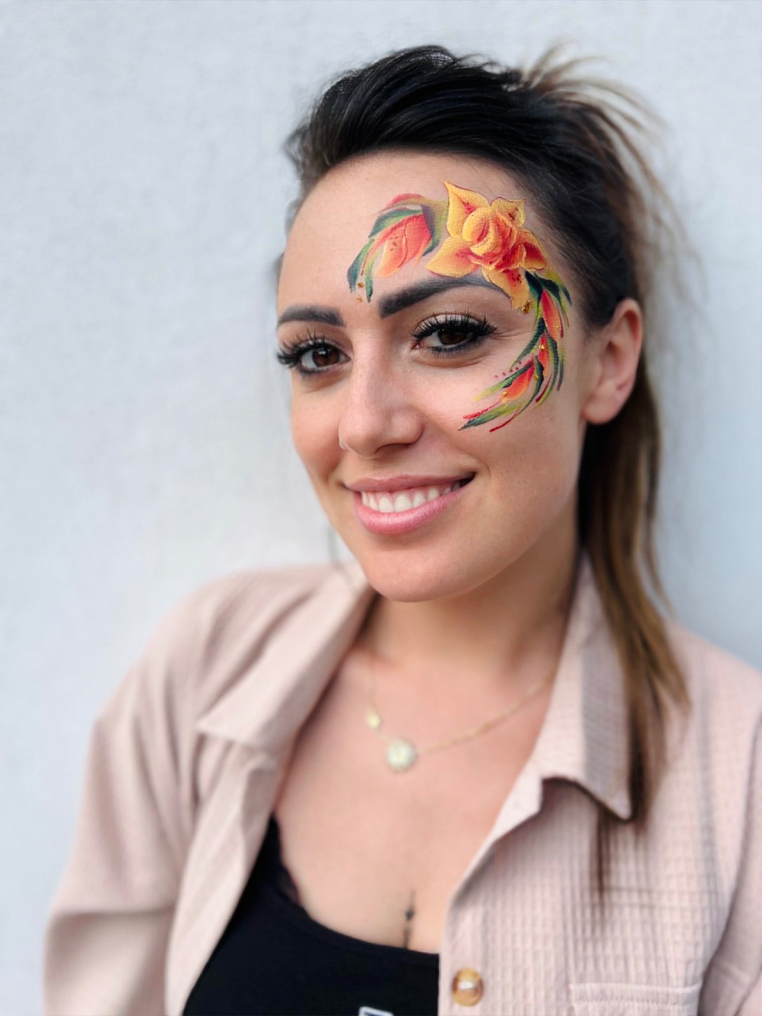 Uśmiechnięta młoda kobieta z profesjonalnym makijażem artystycznym na twarzy przedstawiającym żółto-czerwony kwiat z zielonymi liśćmi, skierowana wprost do widza.