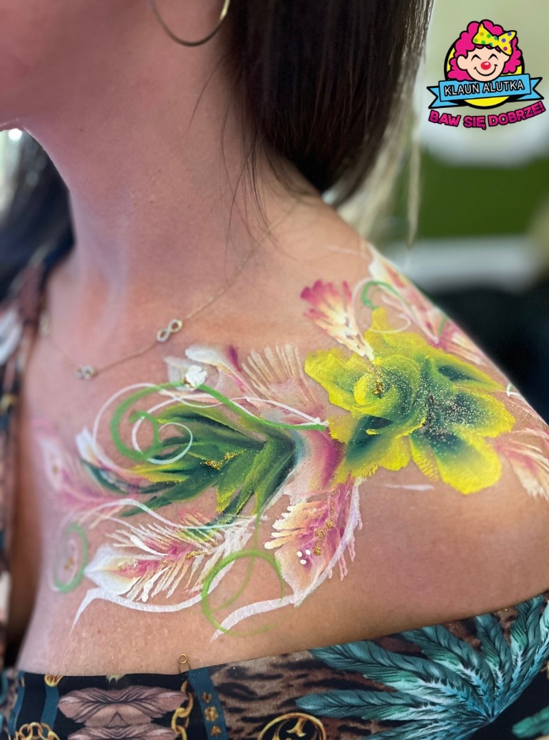 Kobieta z malunkiem na ramieniu i dekolcie przedstawiającym kwiaty i liście w odcieniach zieleni, różu i żółci, z logo firmy Klaun Alutka w prawym górnym rogu.
