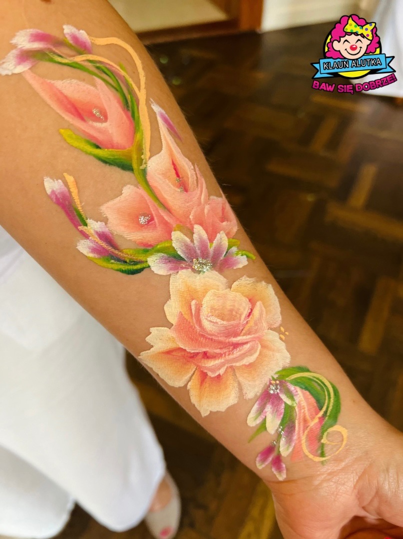 Ręka z artystycznym malunkiem przedstawiającym kompozycję kwiatową w odcieniach różu i zieleni, z dodatkiem srebrnego brokatu, wykonanym na skórze.