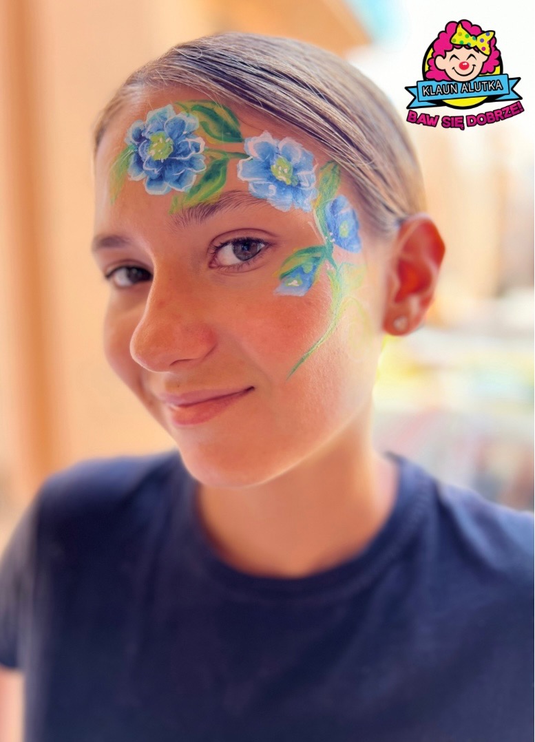 Portret uśmiechniętej dziewczyny z kwiatowym malunkiem na czole i policzku, wykonanym farbami do twarzy w odcieniach niebieskiego i zieleni.
