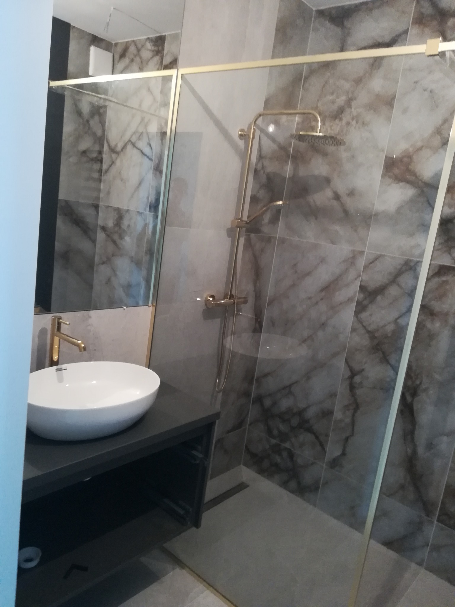 Nowoczesna łazienka z marmurowymi płytkami, złotą armaturą i kabiną prysznicową typu walk-in, umywalka nablatowa na ciemnoszarym blacie.