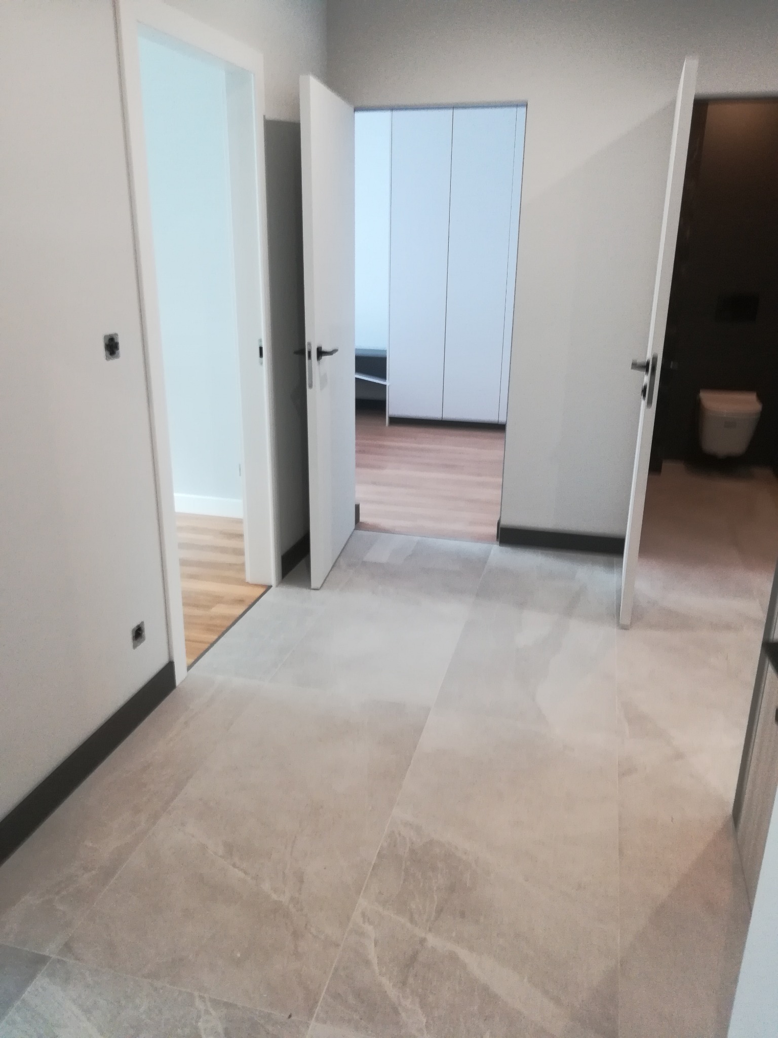 Korytarz z beżową marmurową podłogą, widoczne otwarte białe drzwi do pomieszczeń z drewnianą podłogą i toalety, ciemne listwy przypodłogowe.