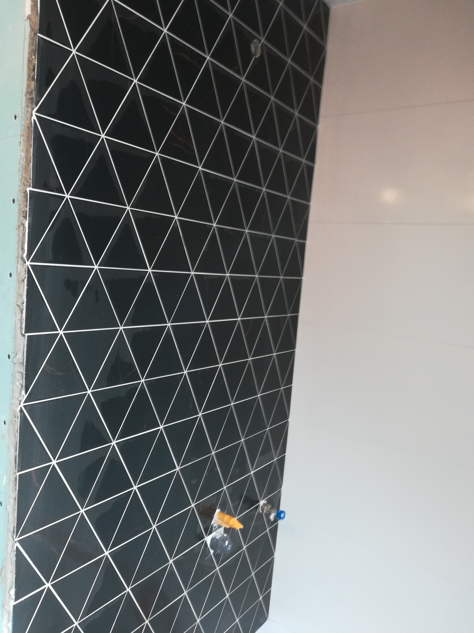 Układanie czarnych, trójkątnych płytek z białymi fugami na ścianie w trakcie prac wykończeniowych, widoczne narzędzia i fragment białej ściany.