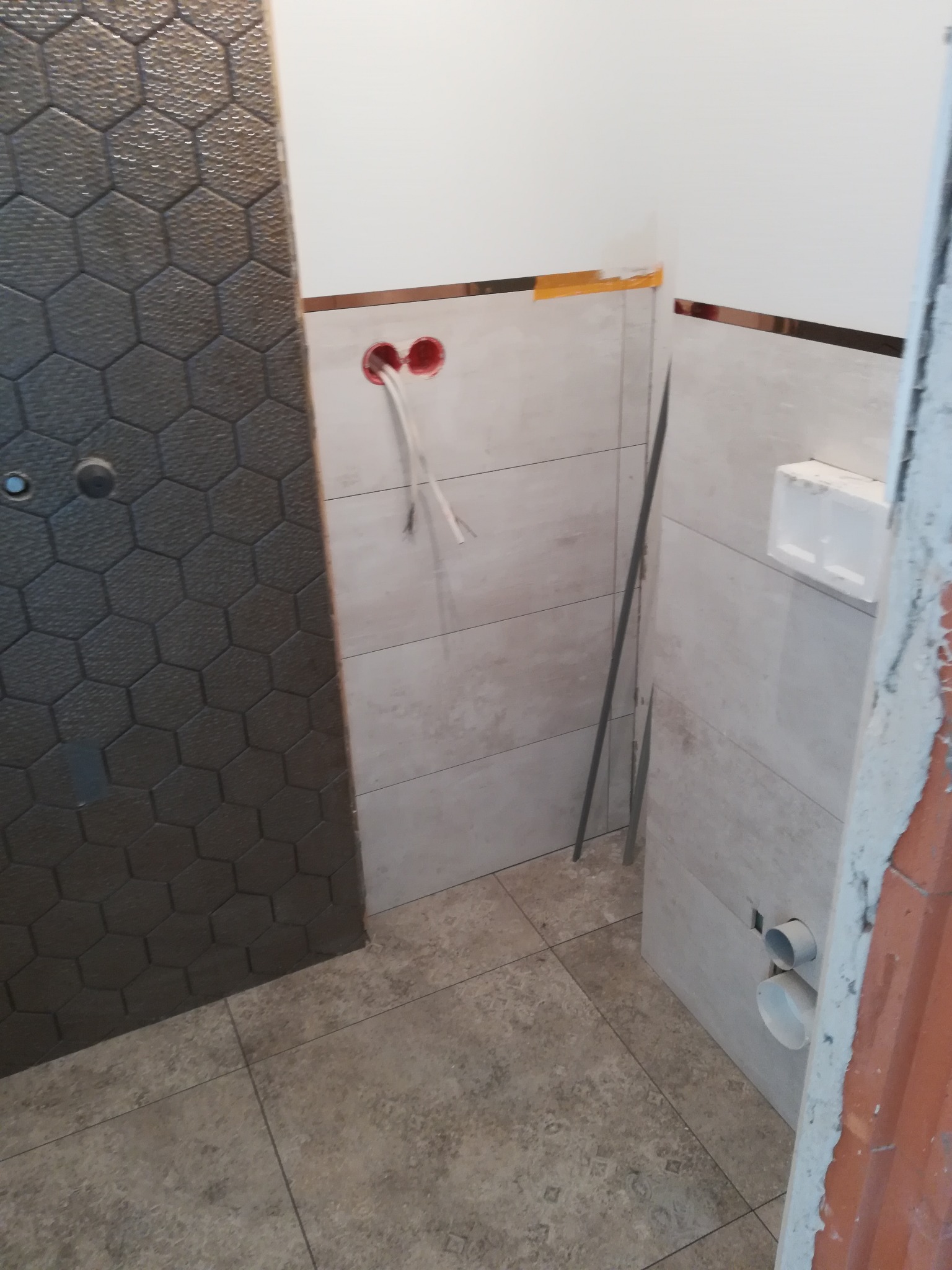 Łazienka w trakcie remontu: położone płytki podłogowe i ścienne w odcieniach beżu i szarości, widoczne wystające kable elektryczne, rury odpływowe, oraz fragment ściany wykończonej heksagonalną...