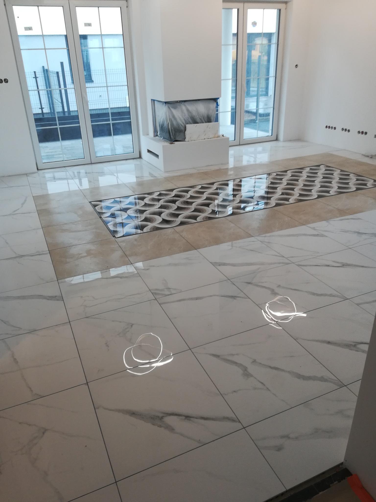 Jasne marmurowe płytki podłogowe z dekoracyjnym wzorem wstawionym centralnie, widoczne duże okna i fragment obudowy kominka zabezpieczonego folią.